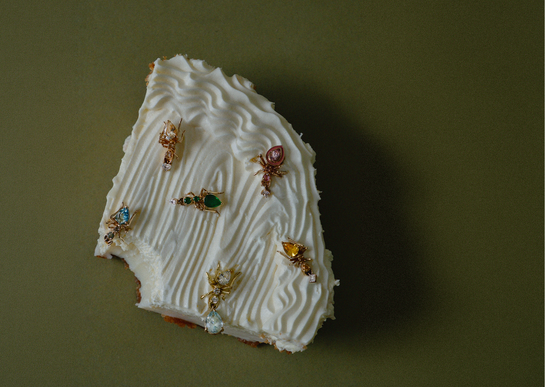 A small irregular white cake slice topped with piped cream and several small ant earring studs by cocoerow fine jewelry
